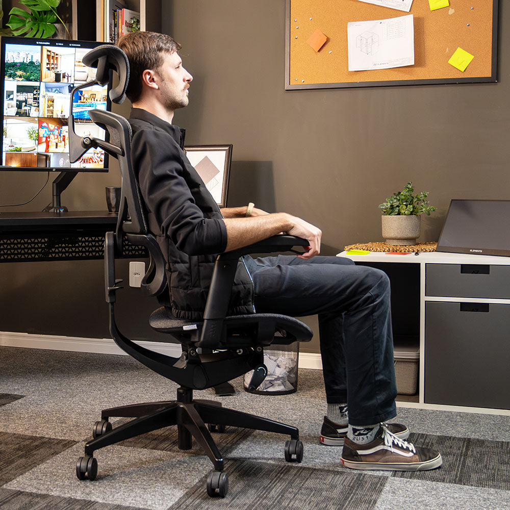 A imagem mostra um homem sentado numa cadeira ergonômica Nemesis num espaço home office organizado com um setup composto de uma mesa de altura, teclado e um monitor