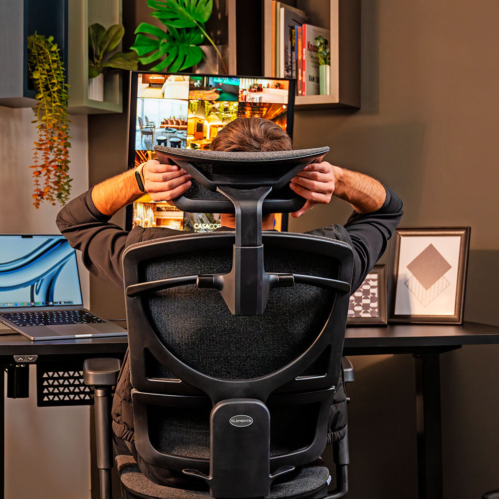 A imagem mostra um homem sentado numa cadeira ergonômica Nemesis ajustando o suporte cervical, num espaço home office organizado com um setup composto de uma mesa de altura, um notebook e um monitor