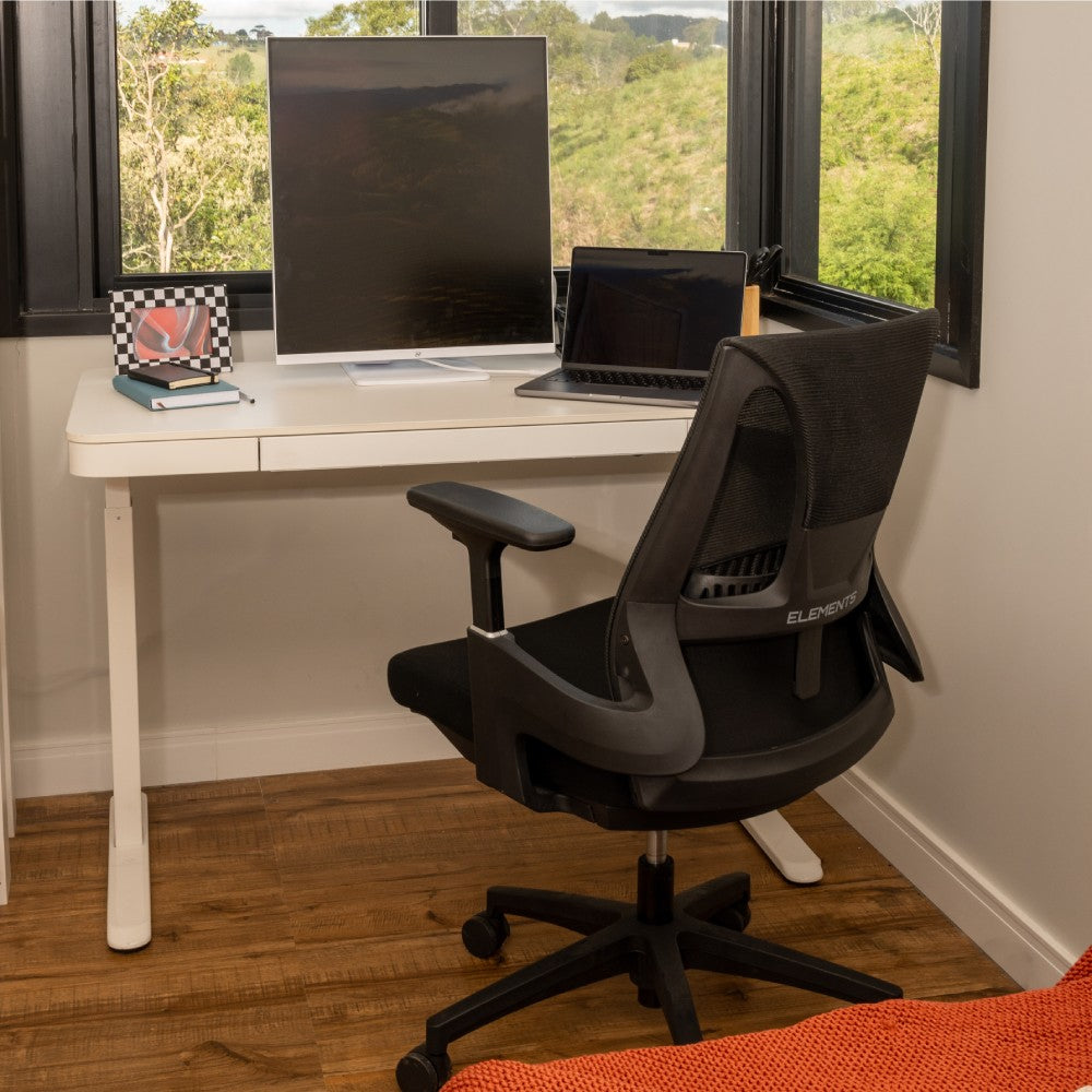 Cadeira ergonômica Elements em um ambiente de home office com mesa branca, monitor, laptop e caderno, em um espaço iluminado com vista para a natureza.