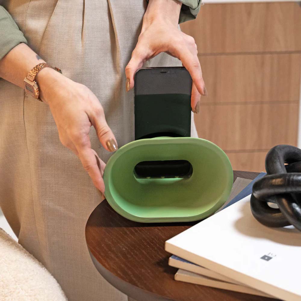 A imagem mostra uma mesa de café de madeira clara com diversos objetos sobre ela. Em destaque, há um amplificador de som Monsoni, e uma moça colocando um smartphone no amplificador.