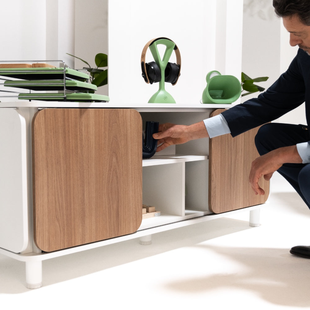 A imagem mostra um homem ajoelhado em frente a um armário Appennini Low, enquanto coloca ou retira um objeto decorativo em uma das prateleiras internas do móvel. O buffet tem portas de madeira com estrutura branca, e sobre ele há itens organizacionais e decorativos, como um suporte verde para fones de ouvido, um organizador de papéis em verde e uma luminária verde.
