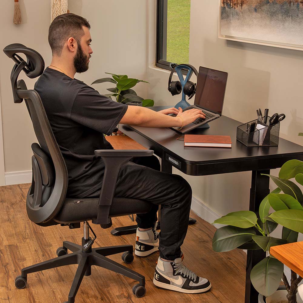 "Imagem de uma pessoa sentada confortavelmente na cadeira ergonômica Astra Pro, trabalhando na mesa Musa em um ambiente acolhedor. A pessoa demonstra boa postura, com o apoio lombar da cadeira contribuindo para o conforto durante a atividade. O cenário exibe uma atmosfera relaxante, ideal para um espaço de trabalho produtivo."