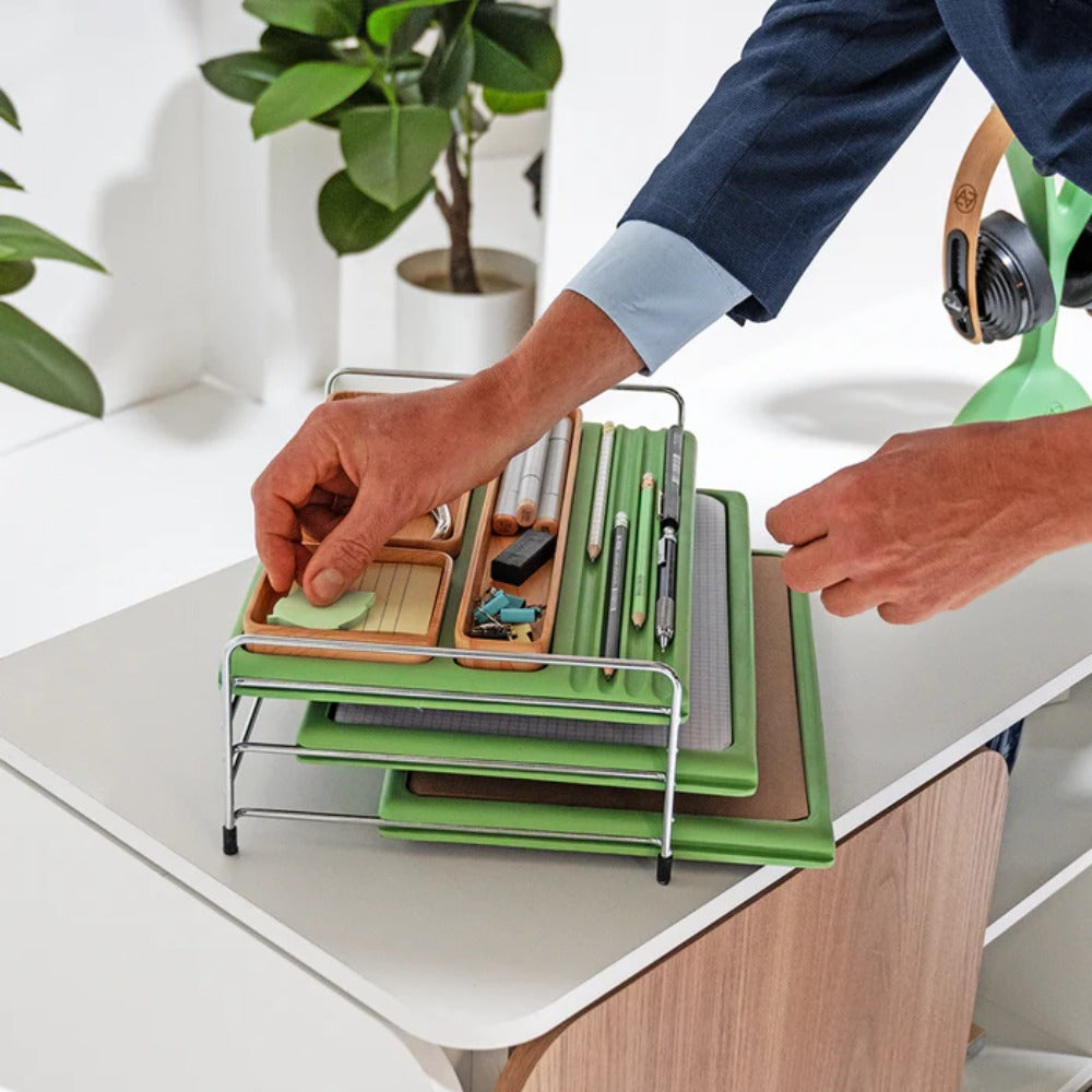 A imagem mostra um homem pegando artigos do conjunto de Bandejas organizadoras Baltea com três prateleiras verdes empilhadas em uma estrutura de metal cromado. No topo, há compartimentos de madeira com objetos de papelaria, como tesoura, perfurador, post-its, clipes de papel, lápis e canetas. O organizador está colocado sobre um armário branco, e ao fundo, há uma planta decorativa que complementa o ambiente.