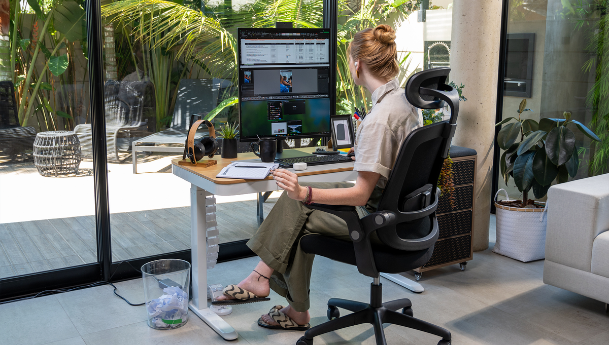 cadeira ergonômica Astra com uma modelo femininia com uma mesa regulagem de altura em uma casa com grandes janelas para um espaço com plantas.