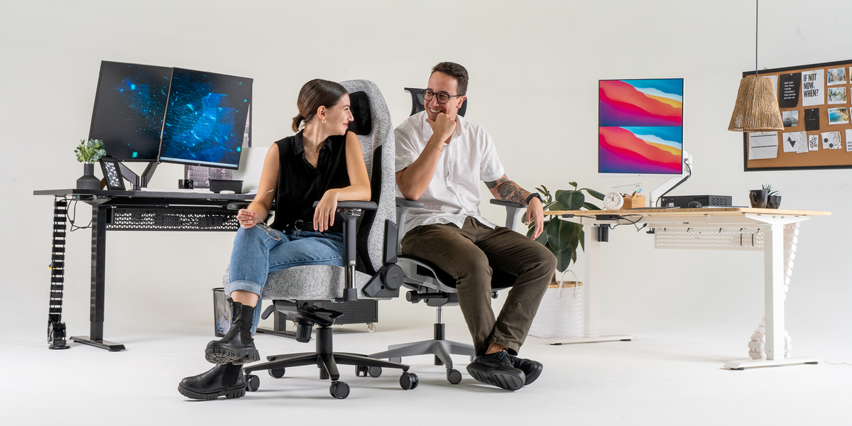 Foto de um casal com duas mesas com regulagem de altura elétrica modelo Mish uma preta e outra branca com uma cadeira modelo Fearless e outra modelo Vertta ambas da marca Elements