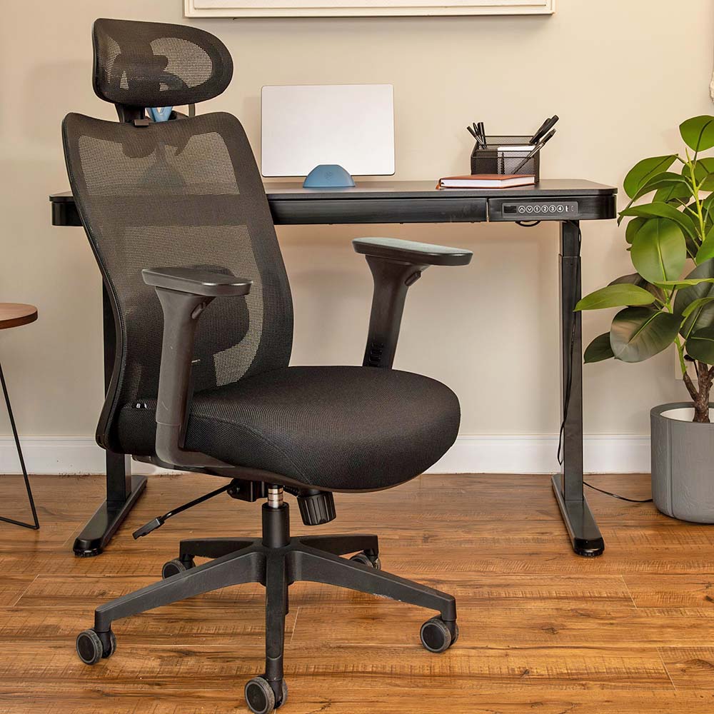 Imagem da cadeira ergonômica Astra Pro em um ambiente de trabalho moderno, com uma mesa ajustável em altura ao fundo e um vaso de planta decorativo. A cadeira destaca-se pelo seu design ergonômico, que promove conforto e saúde postural, enquanto a mesa proporciona flexibilidade no espaço de trabalho, ideal para diferentes necessidades e preferências.