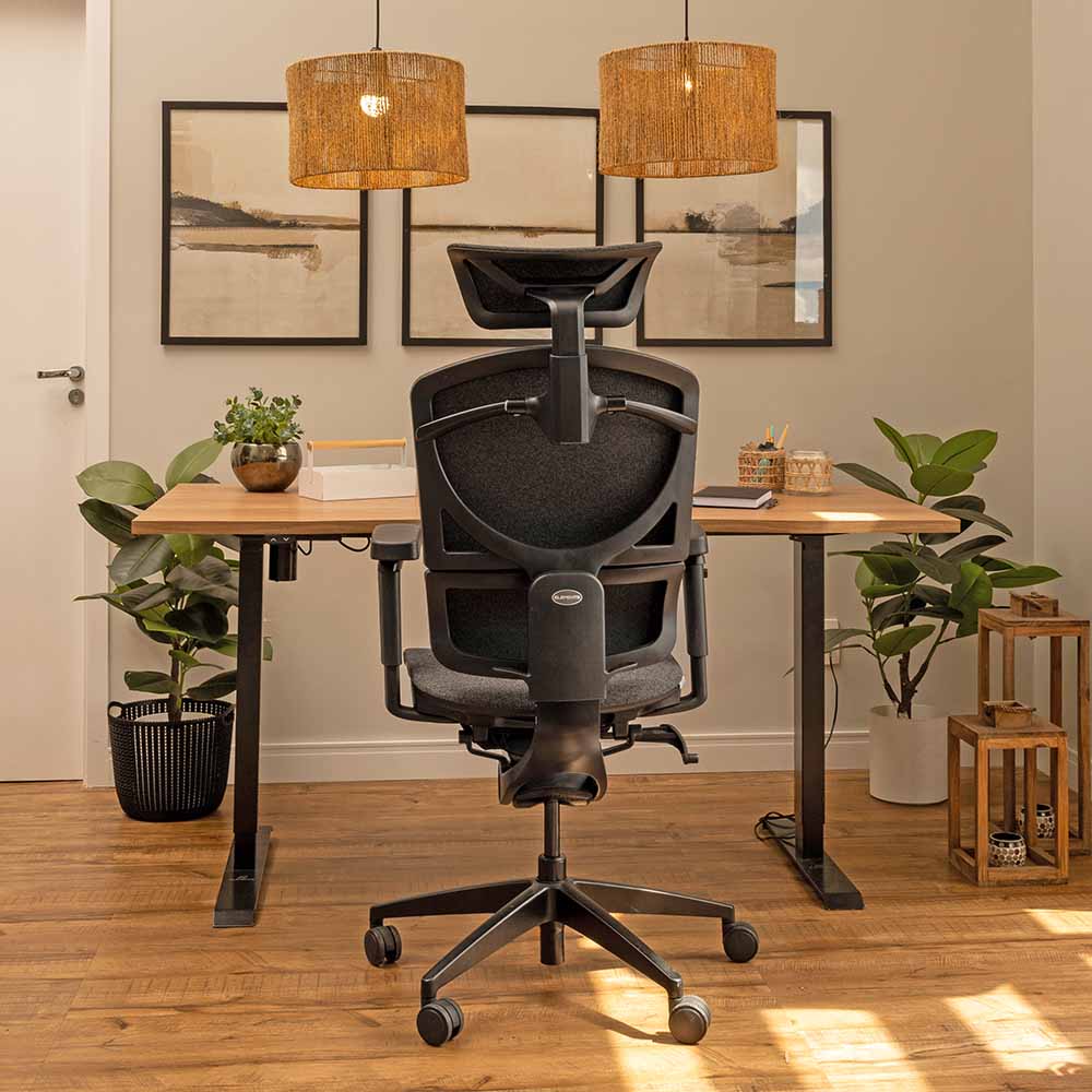 A imagem mostra uma estação de trabalho elegante e acolhedora, com uma cadeira ergonômica de encosto robusto posicionada em frente a uma mesa ajustável em altura Misch com tampo de madeira ergonômico. O ambiente é decorado com plantas e elementos de madeira, além de luminárias pendentes que conferem um toque aconchegante. O piso de madeira e as molduras na parede com arte minimalista completam o espaço, proporcionando um local de trabalho funcional e esteticamente agradável.